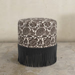 Round upholstered ottoman with floral pattern in chocolate brown and fringe on a concrete floor.