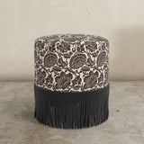 Round upholstered ottoman with floral pattern in chocolate brown and fringe on a concrete floor.