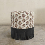 Round upholstered ottoman with pattern and black fringe on a concrete floor.