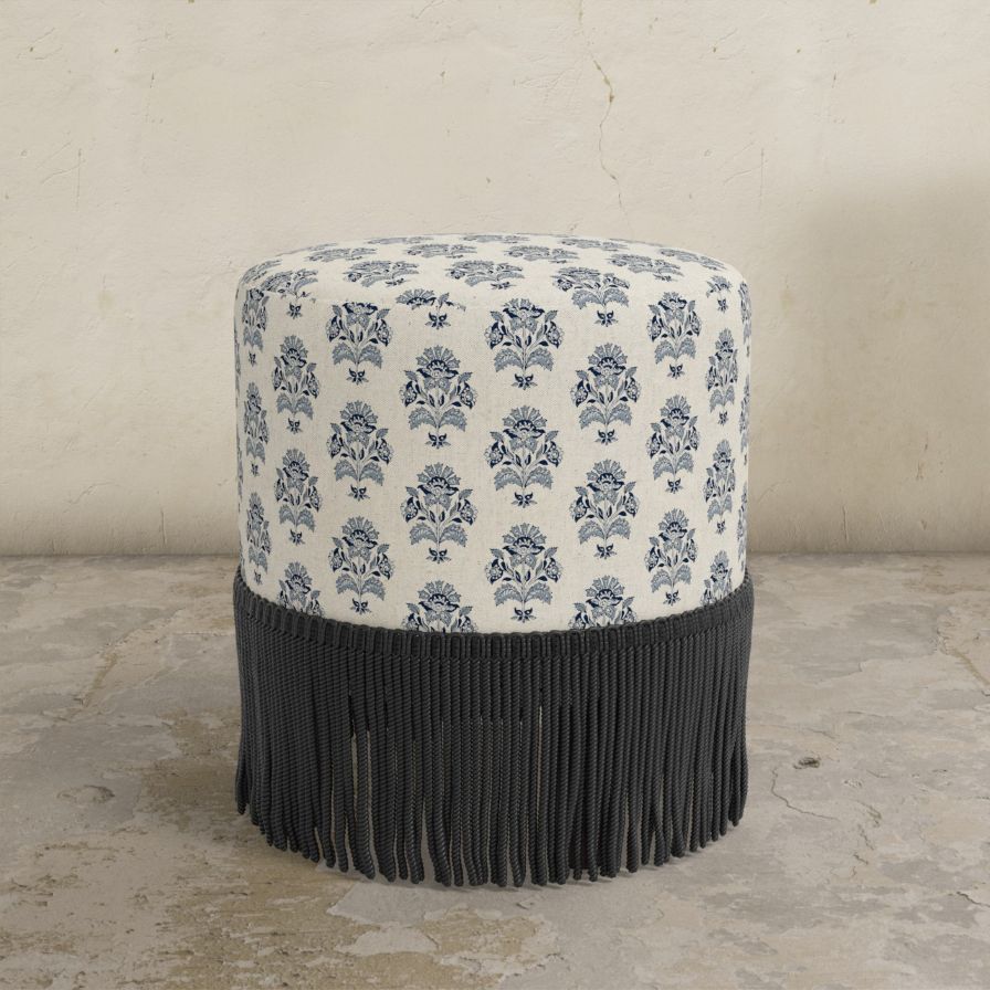 Round upholstered ottoman with pattern and black fringe on a concrete floor.