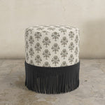 Round upholstered ottoman with pattern top and black fringe bottom on a concrete floor.