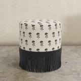 Round upholstered ottoman with pattern and black fringe on a textured floor.
