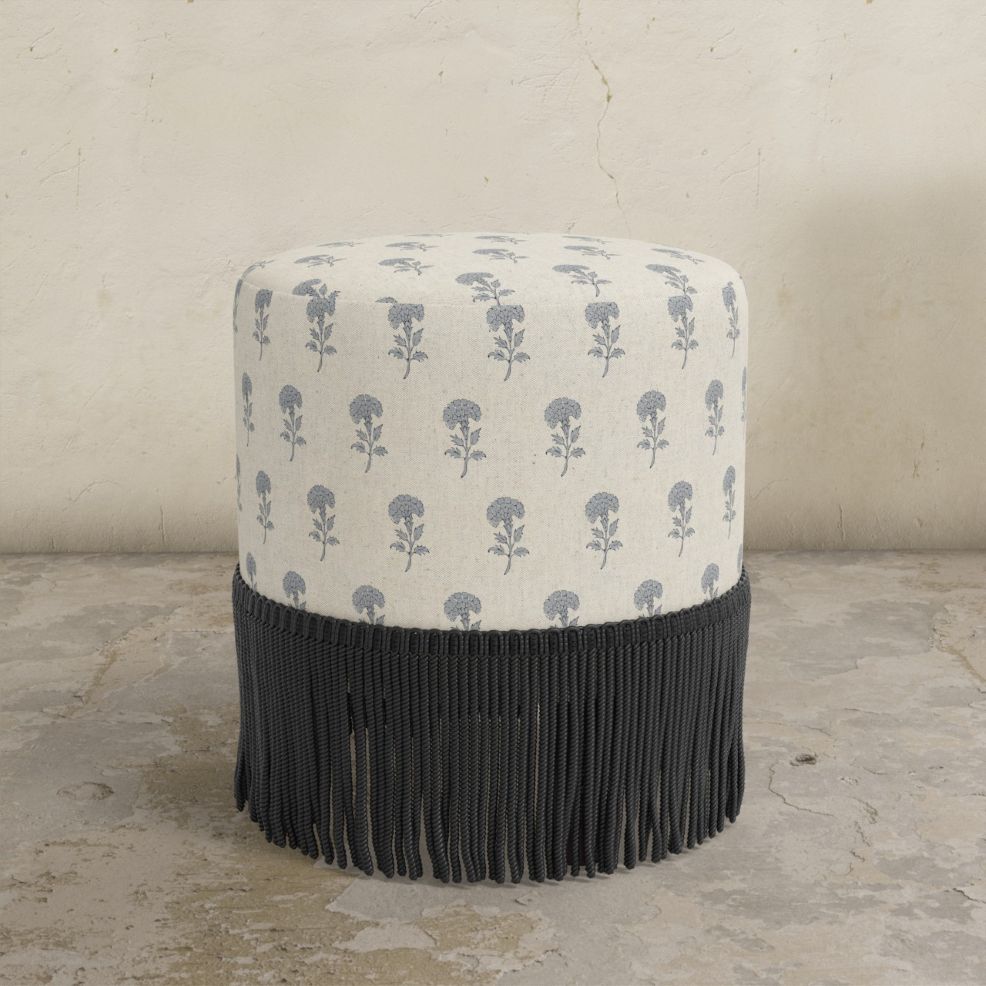 Round upholstered ottoman with blue flower and black fringed bottom on a concrete floor