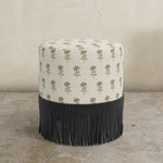 Round upholstered ottoman with patterened top and black fringes on a concrete floor.