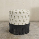 Round upholstered ottoman with patterened top and black fringes on a concrete floor.