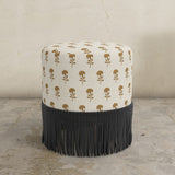 Round upholstered ottoman with patterned top and black fringes on a concrete floor.