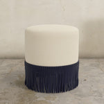 Round natural upholstered ottoman with fringe on a concrete floor.