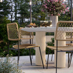 Outdoor dining set featuring a round terrazzo table with fluted pedestal columns and four woven rattan chairs with black metal legs, styled on a patio with a flower arrangement and candles.