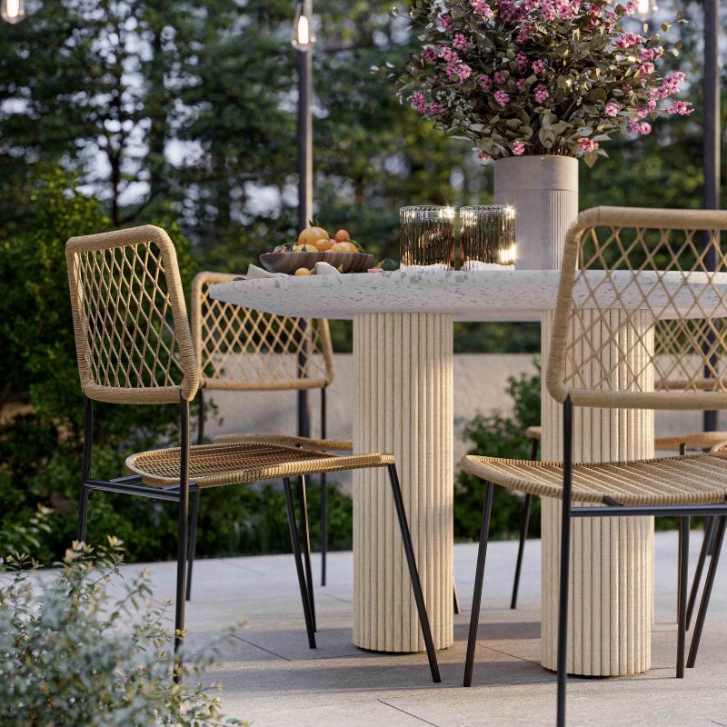 Outdoor dining set featuring a round terrazzo table with fluted pedestal columns and four woven rattan chairs with black metal legs, styled on a patio with a flower arrangement and candles.