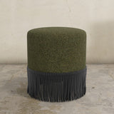 Army green upholstered ottoman with fringe on a concrete floor and light gray wall background