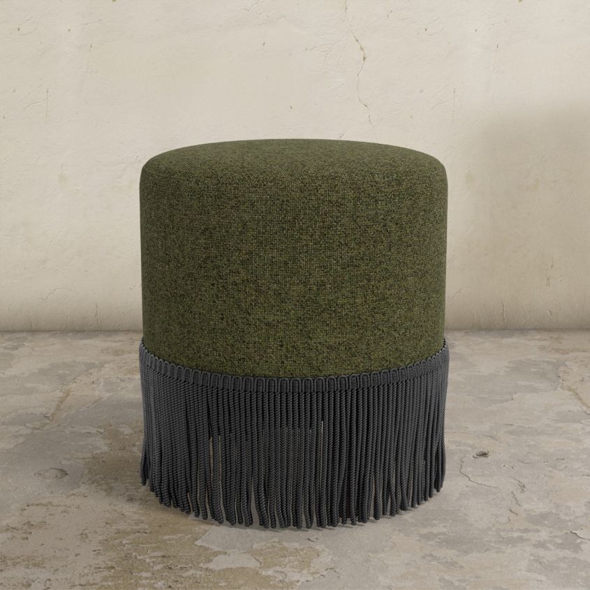Army green upholstered ottoman with fringe on a concrete floor and light gray wall background