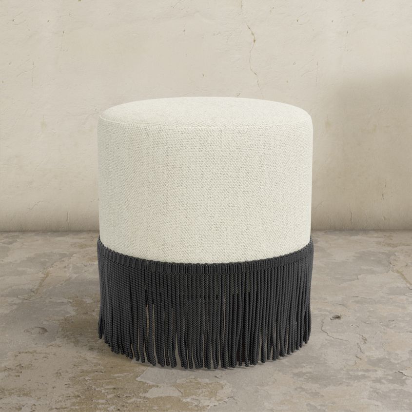Round upholstered ottoman with white top and black fringed bottom on a concrete floor.