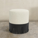 Round upholstered ottoman with white top and black fringed bottom on a concrete floor.