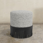 Round ottoman with textured gray top and black fringed bottom on a concrete floor.