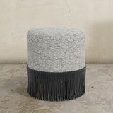 Round ottoman with textured gray top and black fringed bottom on a concrete floor.
