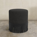 Black ottoman with fringe on a concrete floor and light gray wall background