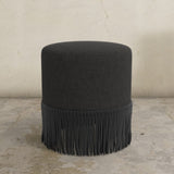 Black ottoman with fringe on a concrete floor and light gray wall background