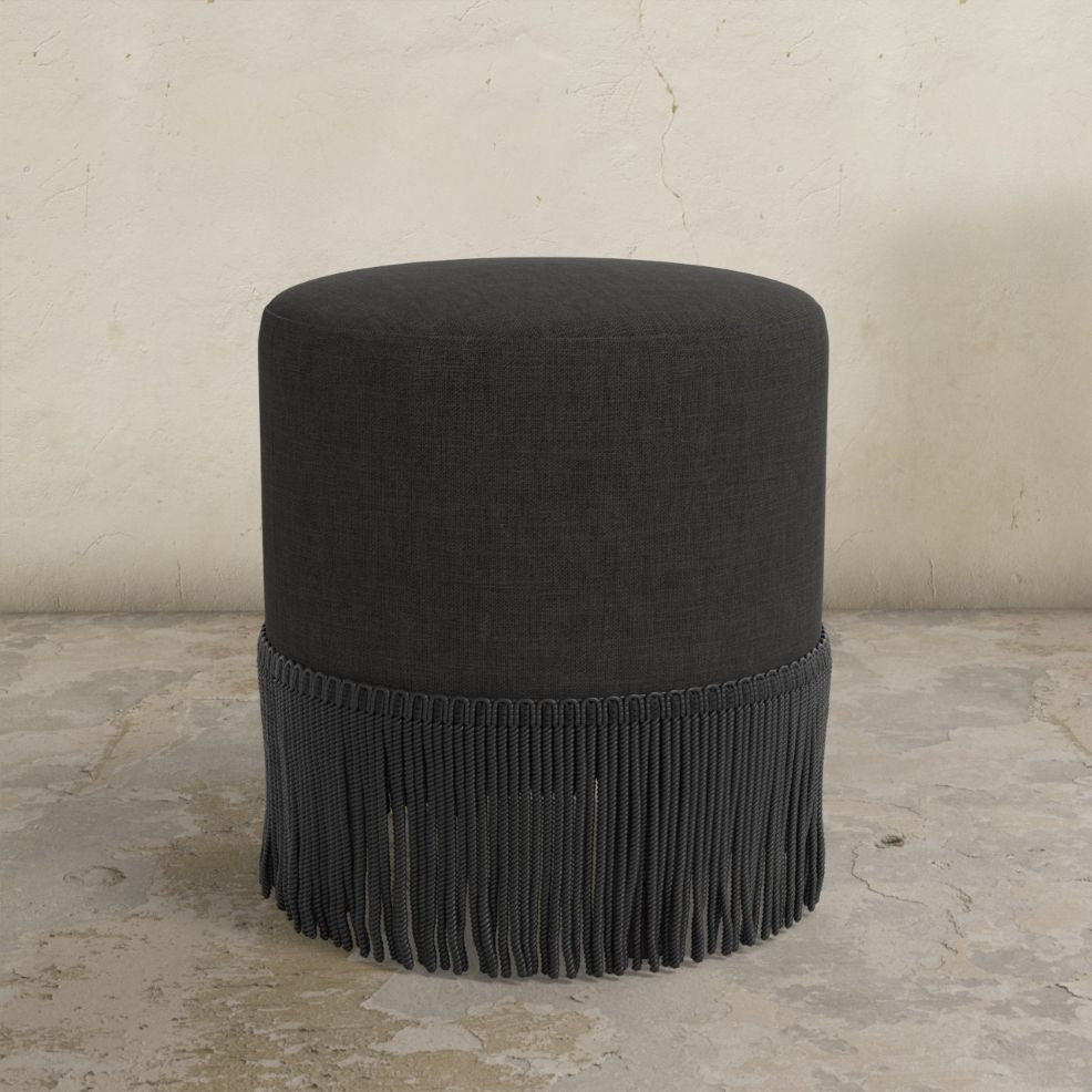 Black ottoman with fringe on a concrete floor and light gray wall background