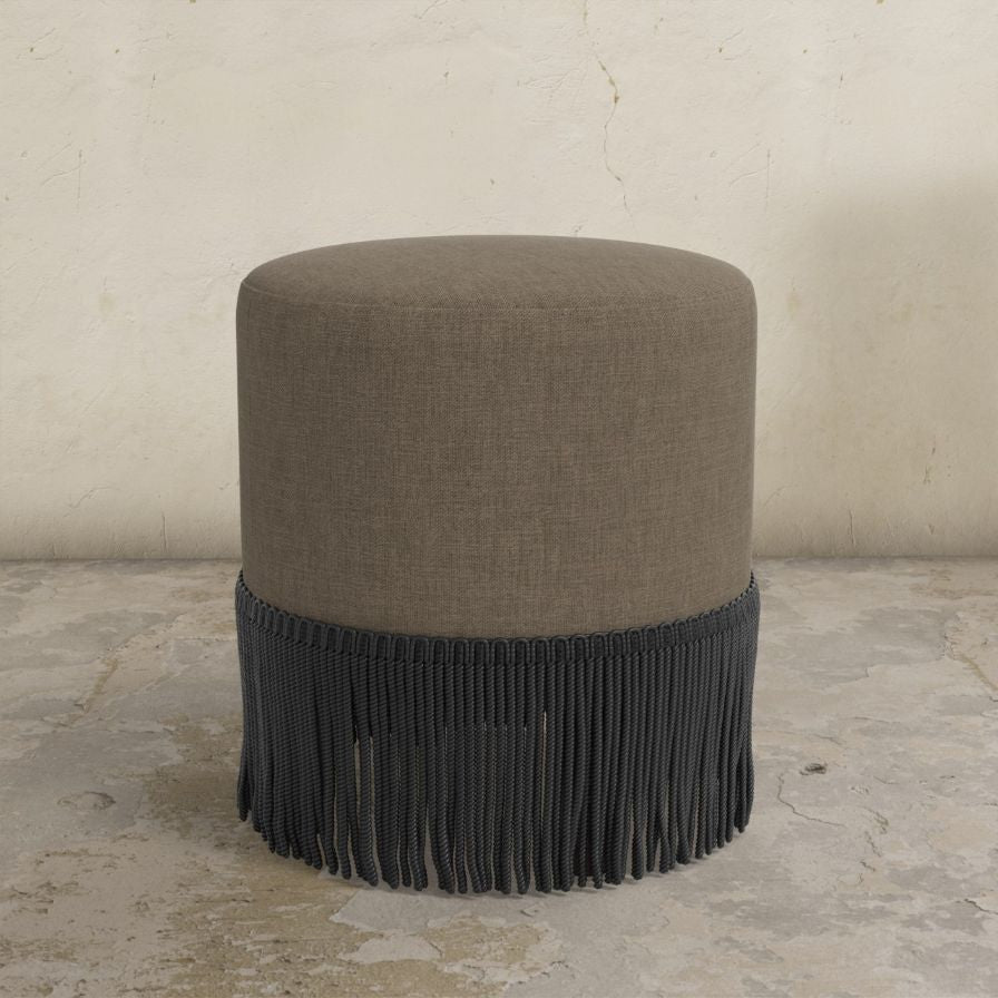 Round upholstered ottoman with fringed bottom on a concrete floor.
