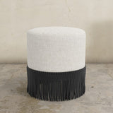 Round upholstered ottoman with white cushion and fringes on a concrete floor.