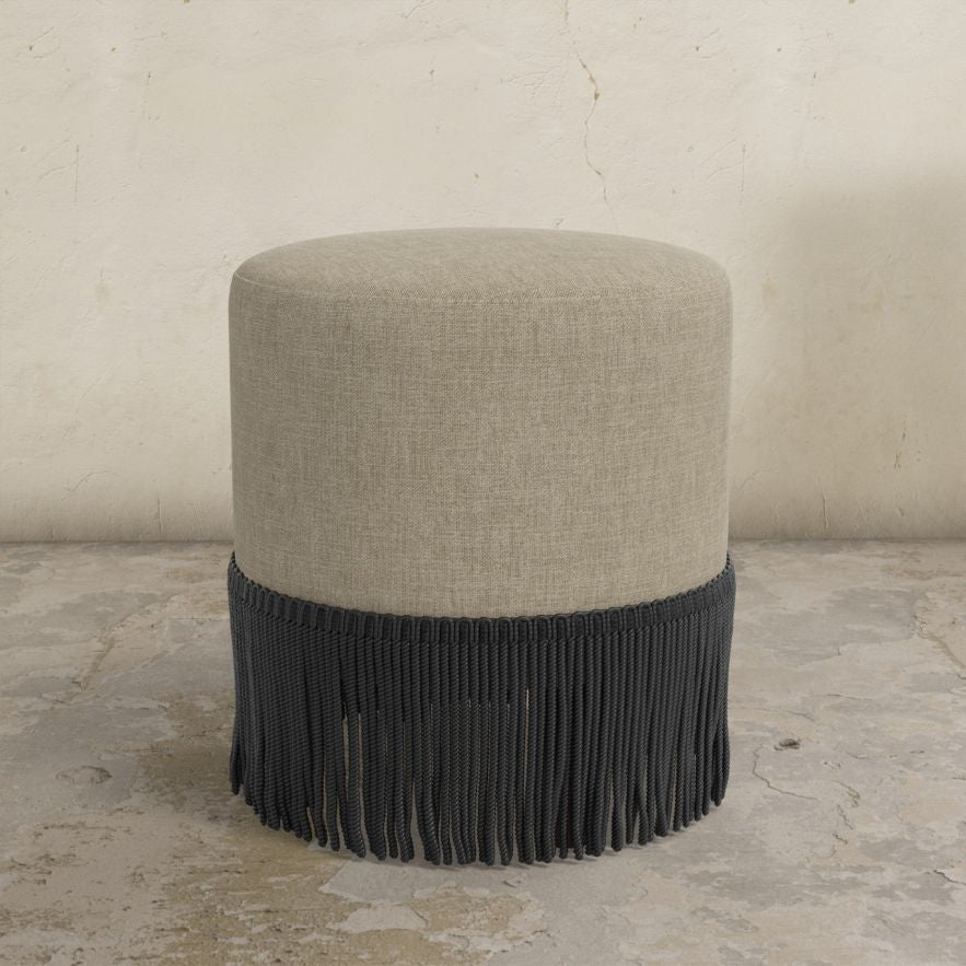 neutral upholstered ottoman with fringe on a concrete floor.