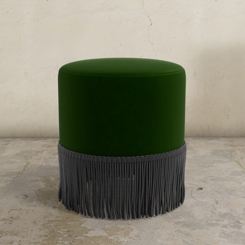 Green upholstered ottoman with gray fringe on a textured floor and light-colored wall background