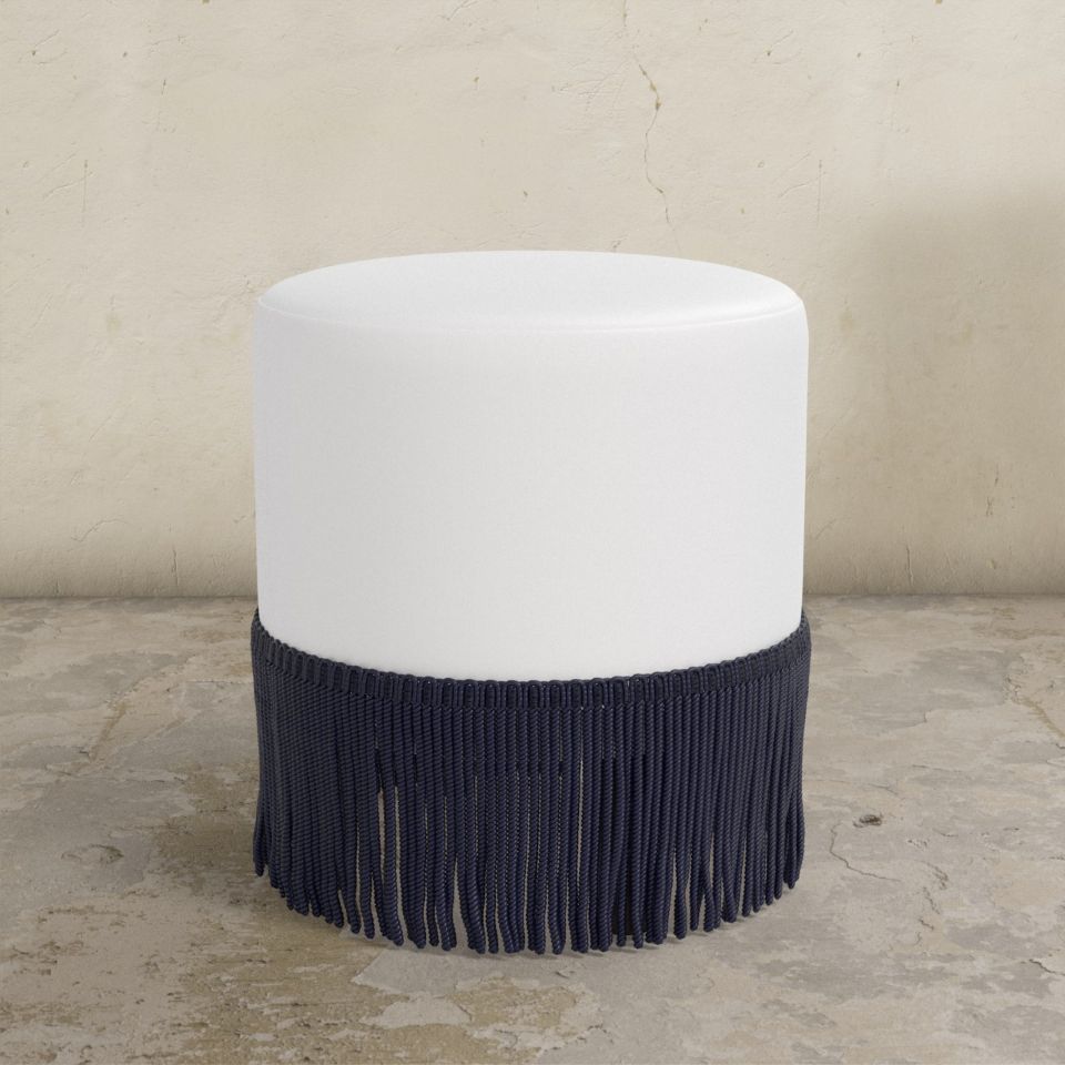 White upholstered ottoman with black fringes on a beige floor.