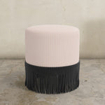 Round blush pink striped upholstered ottoman with fringes on a concrete floor.