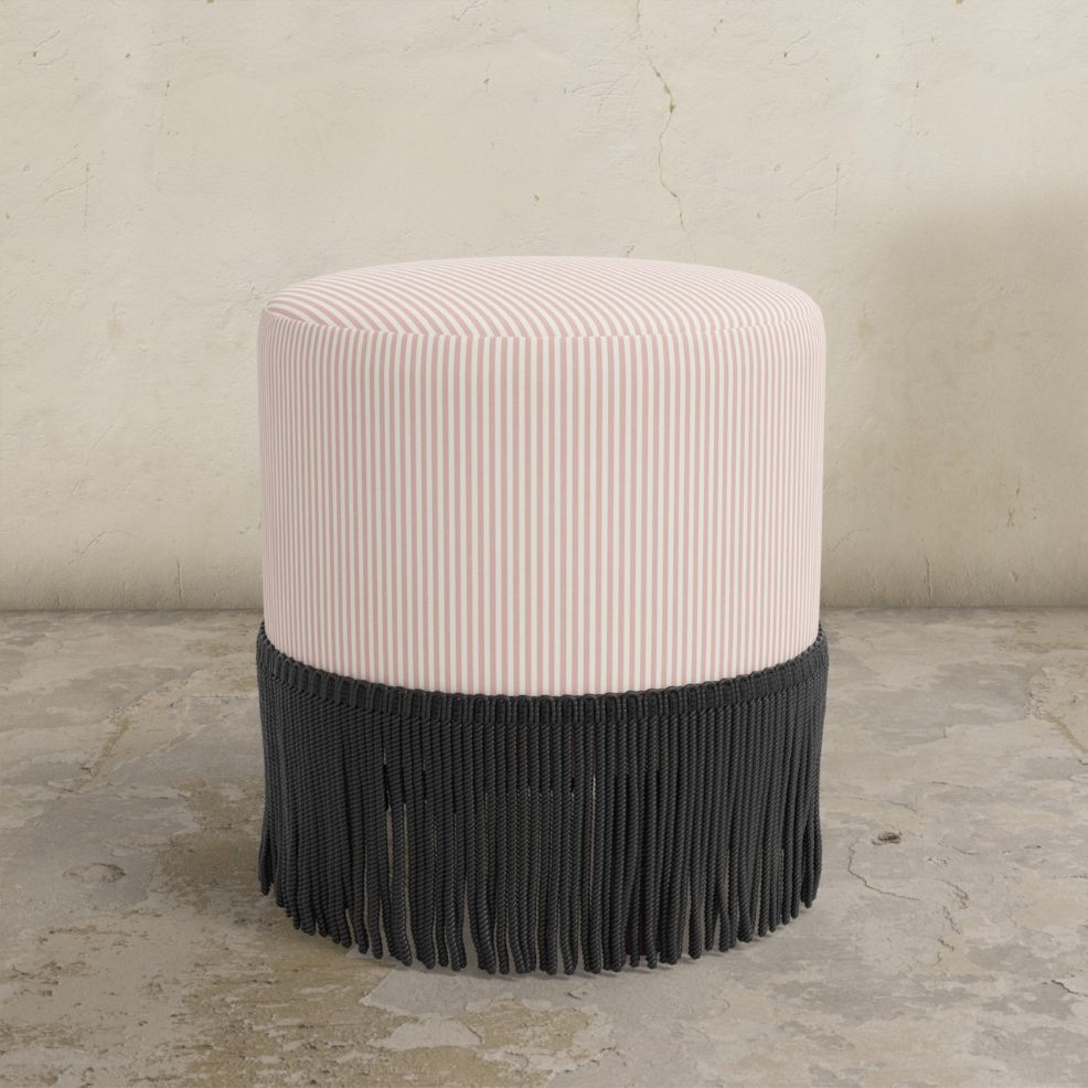 Round blush pink striped upholstered ottoman with fringes on a concrete floor.