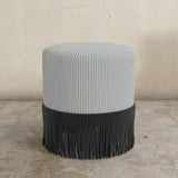 Round upholstered ottoman with blue and white striped top and fringed bottom on a concrete floor.