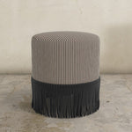 chocolate brown striped upholstered  ottoman with fringe on a concrete floor.