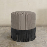 chocolate brown striped upholstered  ottoman with fringe on a concrete floor.