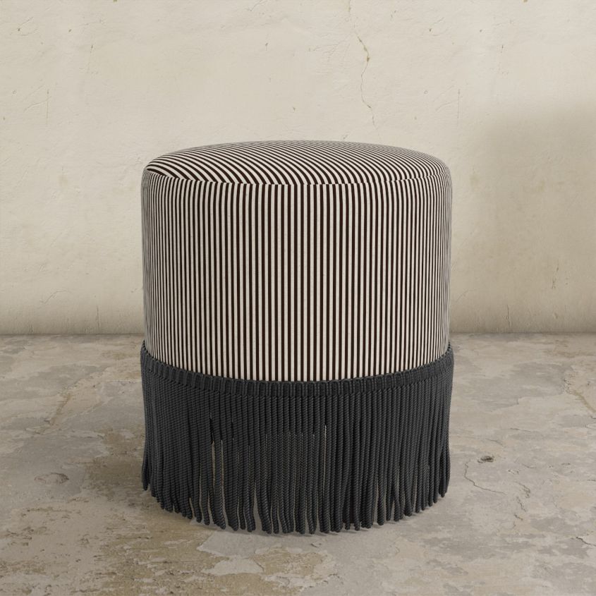 chocolate brown striped upholstered  ottoman with fringe on a concrete floor.