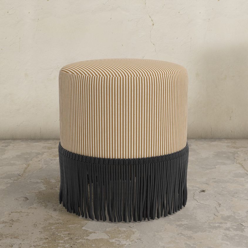 Striped upholstered ottoman with black fringe on a concrete floor.