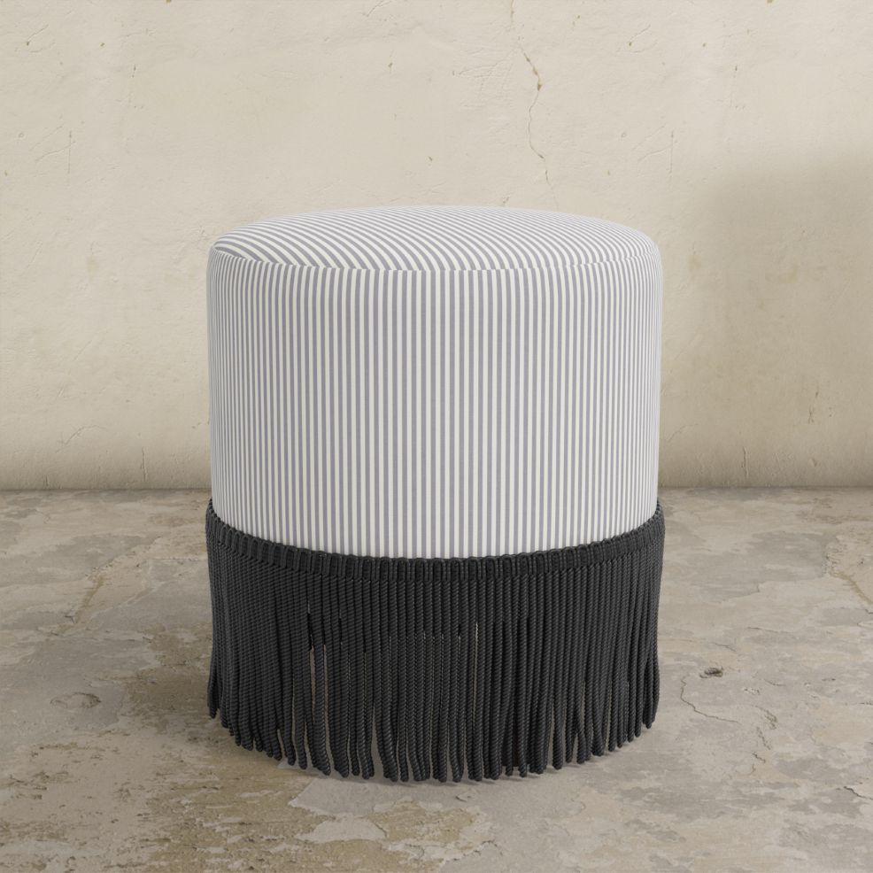 Striped upholstered ottoman with fringe on a concrete floor.
