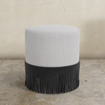 Striped upholstered ottoman with fringe on a concrete floor.