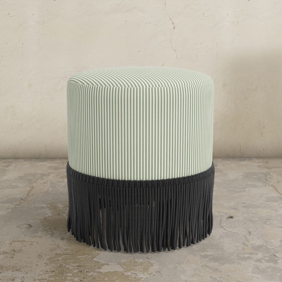 Moss green striped upholstered ottoman with fringe on a concrete floor.