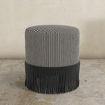 Black striped upholstered ottoman with fringes on a concrete floor against a textured wall.