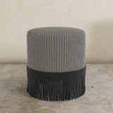 Black striped upholstered ottoman with fringes on a concrete floor against a textured wall.