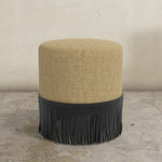 Round upholstered ottoman with beige top and black fringes on a stone floor.