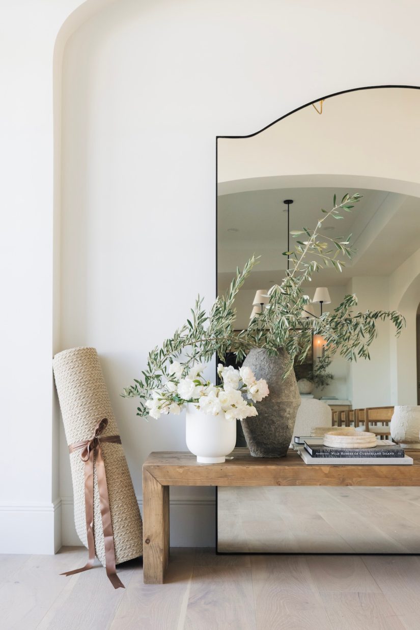 The Ruskin reclaimed pine wood bench styled with a vase of greenery and placed against a white wall, with a floor mirror reflecting the scene.
