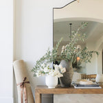 The Ruskin reclaimed pine wood bench styled with a vase of greenery and placed against a white wall, with a floor mirror reflecting the scene.