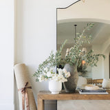 The Ruskin reclaimed pine wood bench styled with a vase of greenery and placed against a white wall, with a floor mirror reflecting the scene.