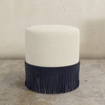 Round upholstered ottoman with white top and navy fringes on a concrete floor.