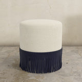 Round upholstered ottoman with white top and navy fringes on a concrete floor.