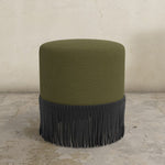 Green upholstered ottoman with black fringe on a stone floor.