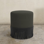Black fringed uphosltered ottoman on a stone floor with a neutral wall background