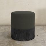 Black fringed uphosltered ottoman on a stone floor with a neutral wall background