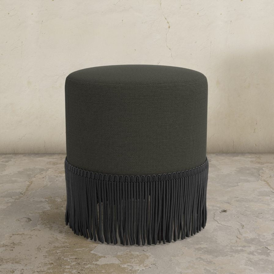 Black fringed uphosltered ottoman on a stone floor with a neutral wall background