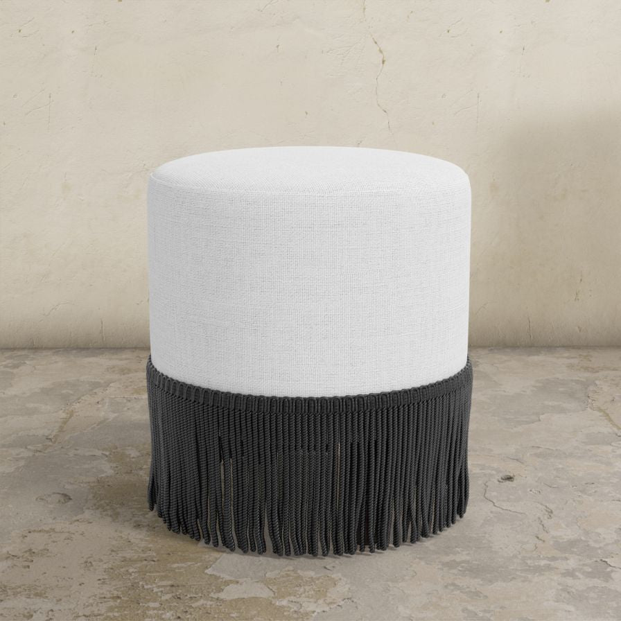 Round upholstered ottoman with white top and black fringed bottom on a textured floor.